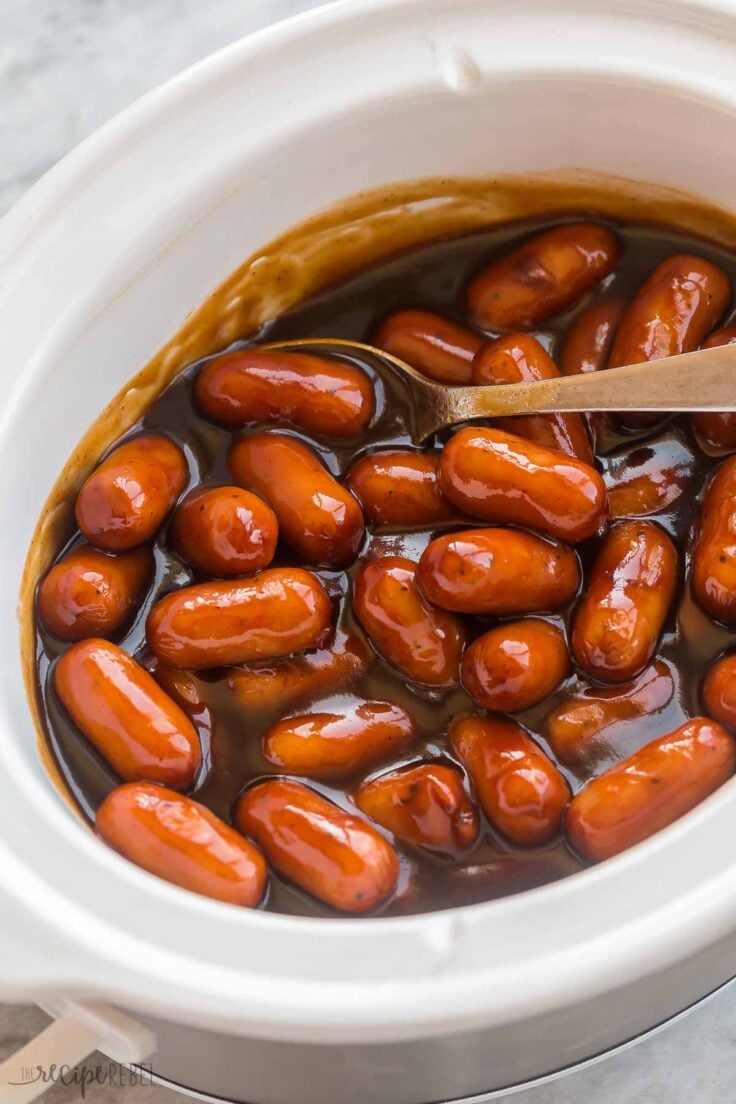 This Little Smokies recipe is the best throw-and-go appetizer or meal! Mini pork sausages are smothered in a sweet, tangy, slightly spicy BBQ brown sugar sauce, then warmed in the slow cooker! #crockpot #slowcooker #recipes | appetizer recipe | easy appetizer | crockpot appetizer | crockpot smokies | bbbq smokies | new years | easy recipe metal spoon scooping lil smokies from the crockpot