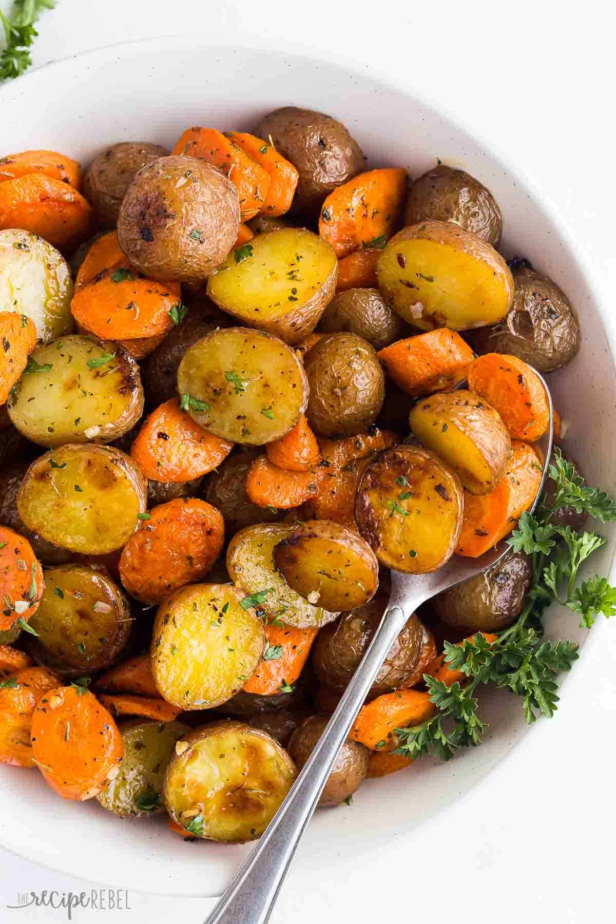 close up image of roasted potatoes and carrots in a white bowl with a spoon.