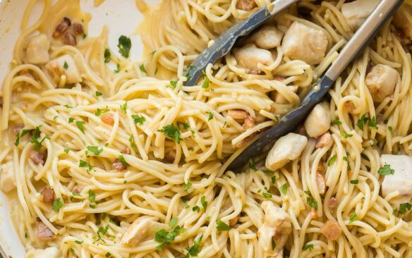 overhead image of chicken carbonara in white skillet with tongs and fresh parsley