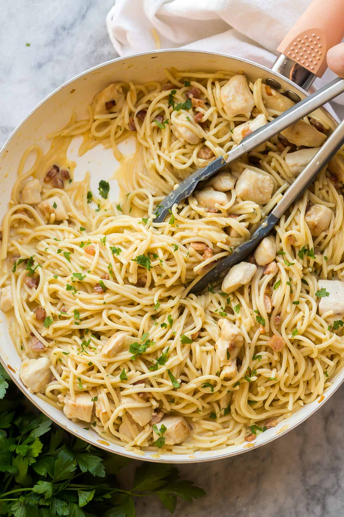 overhead image of chicken carbonara in white skillet with tongs and fresh parsley