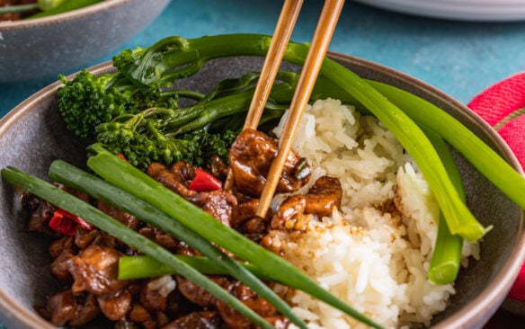 Chopsticks are used to pick up Shanghai chicken served in a bowl with rice and broccolini.
