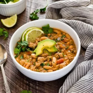 A bowl full of white bean and turkey chili topped with avocado and lime slices.