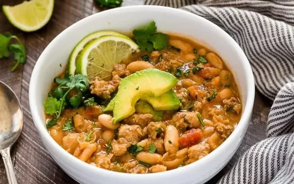 A bowl full of white bean and turkey chili topped with avocado and lime slices.
