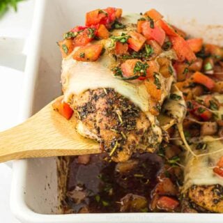 A wooden serving spoon scoops a bruschetta chicken breast out of a baking dish.