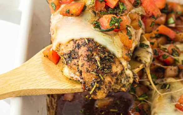 A wooden serving spoon scoops a bruschetta chicken breast out of a baking dish.