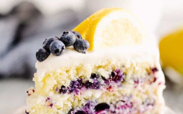 A slice of blueberry lemon cake on a plate, topped with a lemon wedge and fresh blueberries.