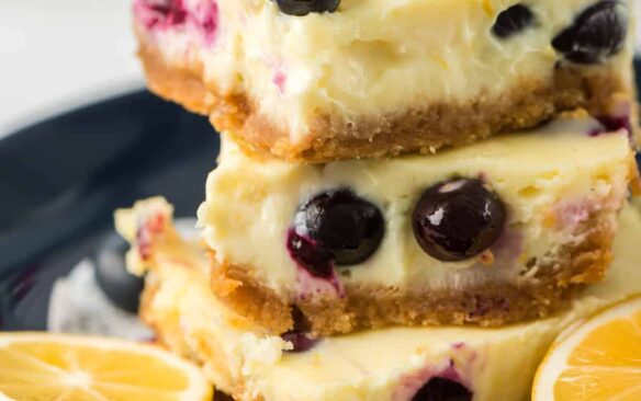 A stack of 3 lemon blueberry cheesecake bars on a plate, surrounded by lemon slices.