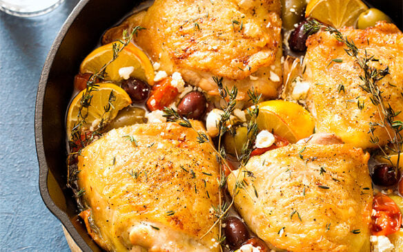 Mediterranean roasted chicken thighs in a skillet with tomatoes, olives, and feta cheese.