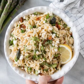 A hand holding a bowl of asparagus and sun dried tomato pasta salad.