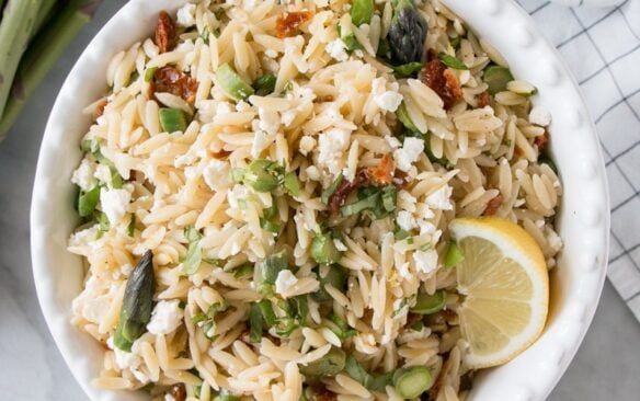 A hand holding a bowl of asparagus and sun dried tomato pasta salad.