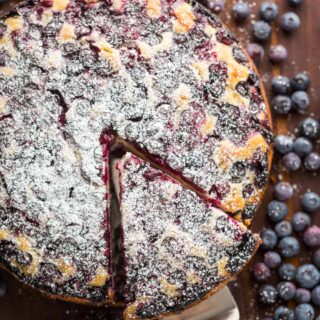 Top view of a blueberry lemon cake with a slice being lifted from it, surrounded by scattered fresh blueberries.