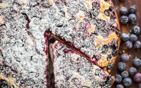 Top view of a blueberry lemon cake with a slice being lifted from it, surrounded by scattered fresh blueberries.