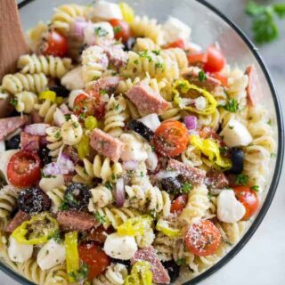 A clear salad bowl full of Italian pasta salad with a wooden spoon.