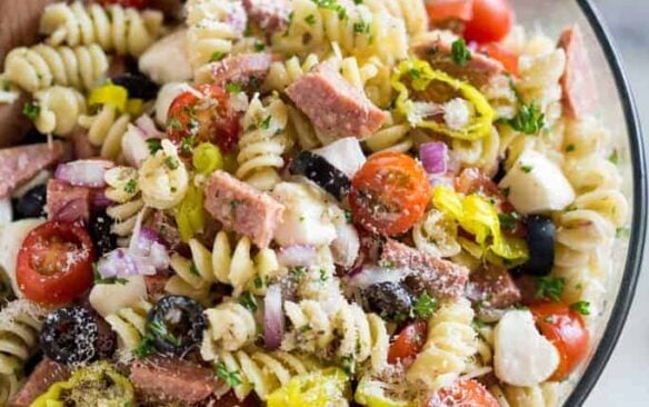 A clear salad bowl full of Italian pasta salad with a wooden spoon.