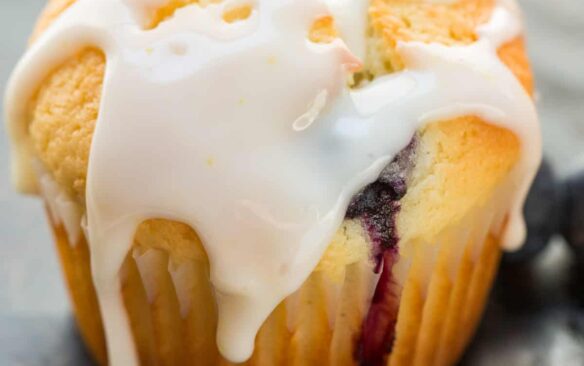 A lemon blueberry muffin topped with glaze, next to fresh blueberries.
