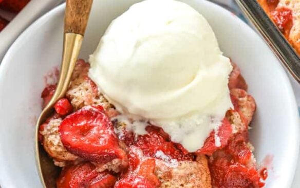 Strawberry rhubarb cobbler in a bowl topped with vanilla ice cream, next to a spoon.