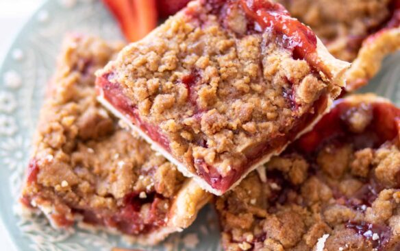 A stack of rhubarb crumb bars on a plate next to fresh sliced strawberries.