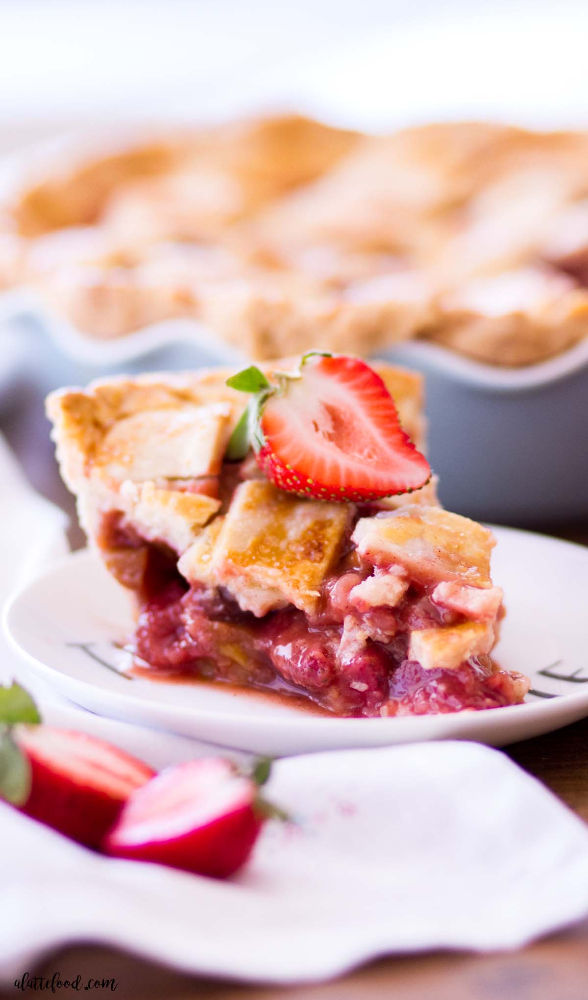 A slice of strawberry rhubarb pie on a plate, with the full pie in the background.