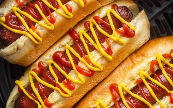 close up image of air fryer hot dogs in air fryer basket with ketchup and mustard