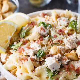 A white bowl of chicken Caesar pasta salad garnished with bacon bits and lemon wedges.