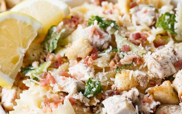 A white bowl of chicken Caesar pasta salad garnished with bacon bits and lemon wedges.