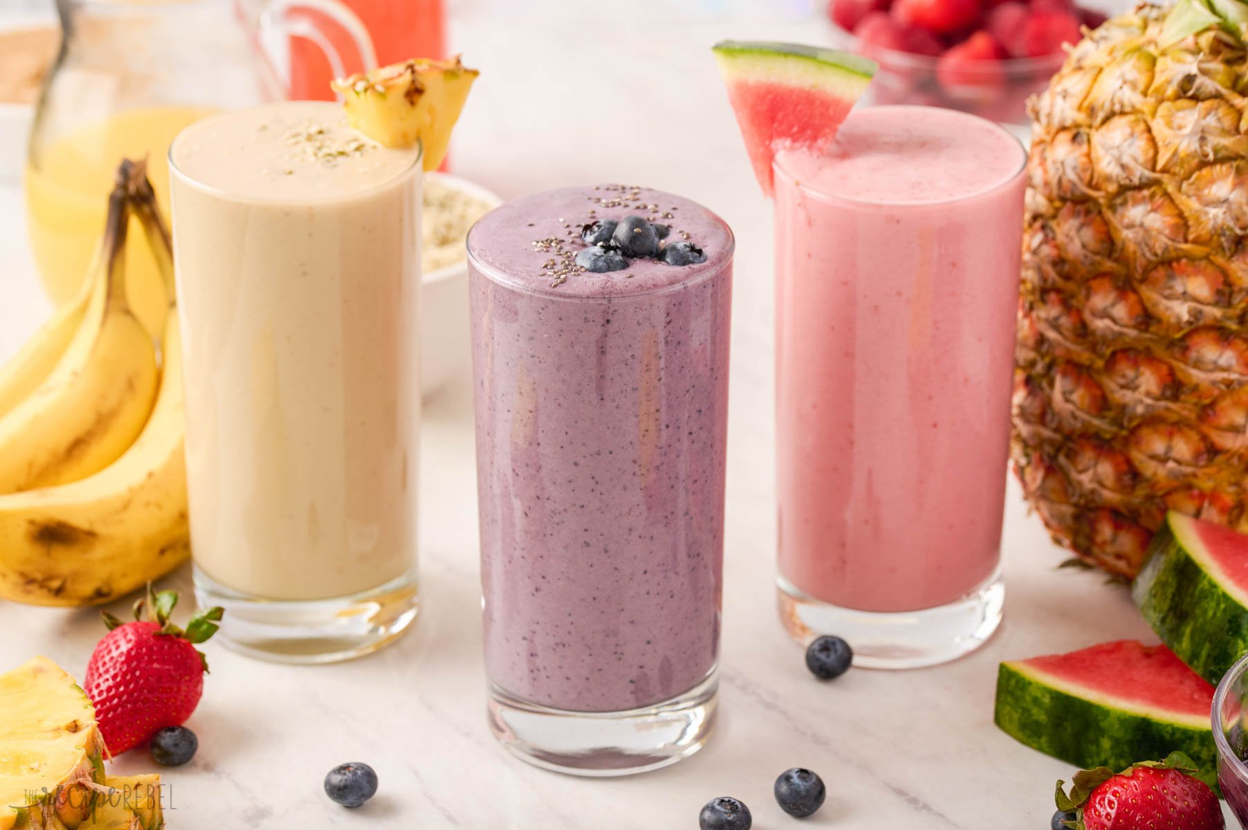 three different smoothies in glasses with a variety of fruit in the background