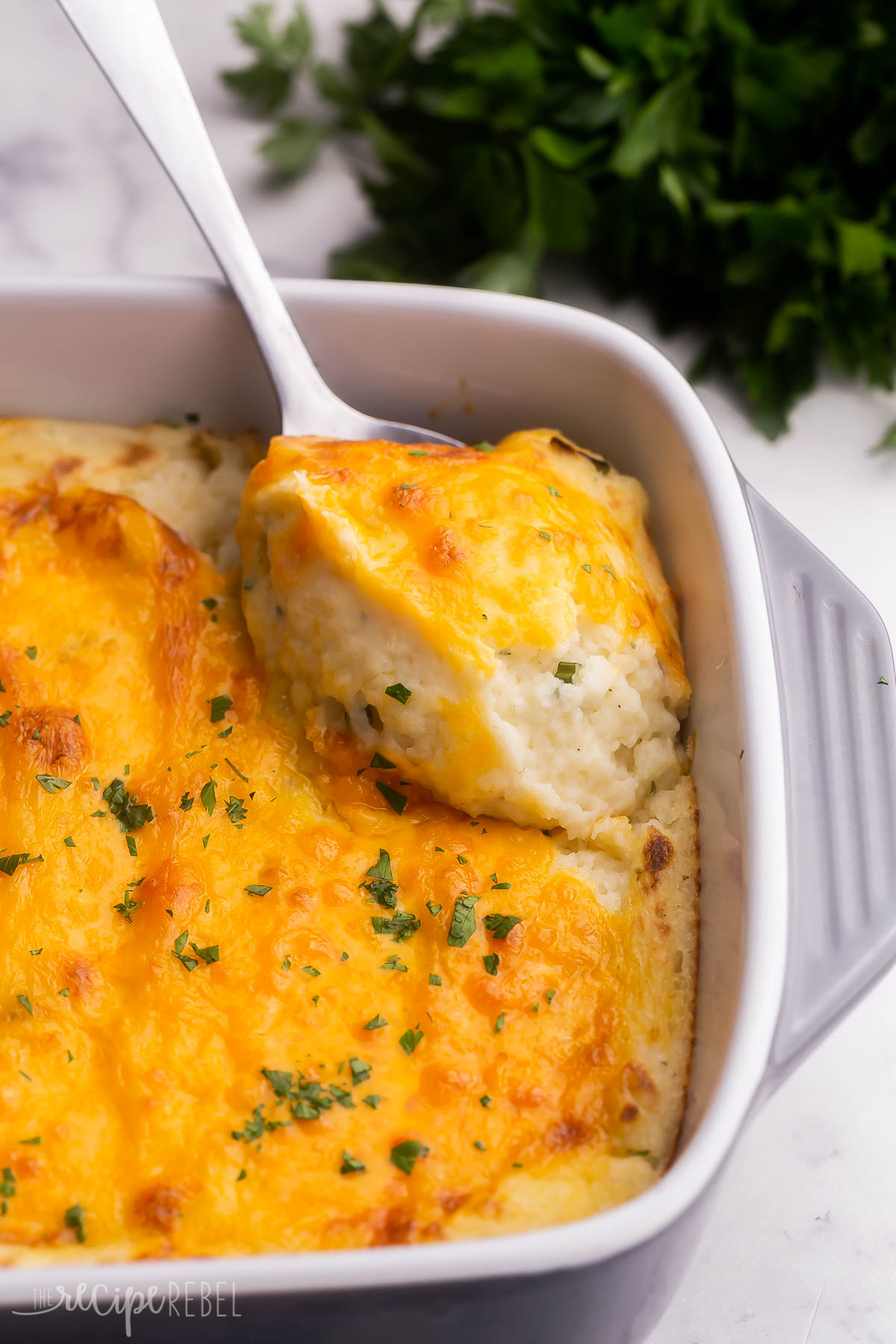 scooping cheesy mashed potatoes out of grey baking dish.