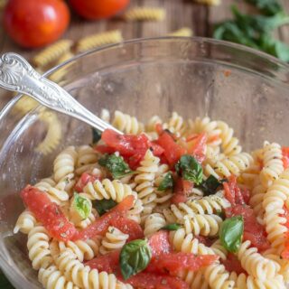 Warm tomato pasta salad in a clear glass bowl with a silver spoon.