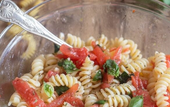 Warm tomato pasta salad in a clear glass bowl with a silver spoon.