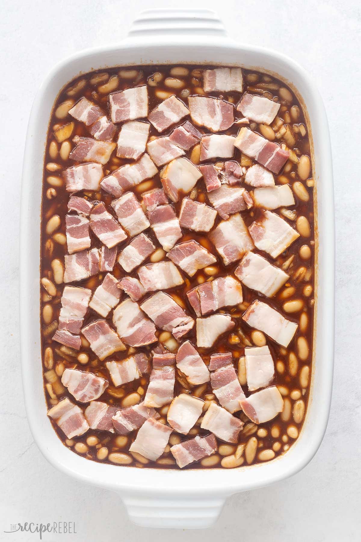 raw pieces of bacon on top of beans in baking dish.
