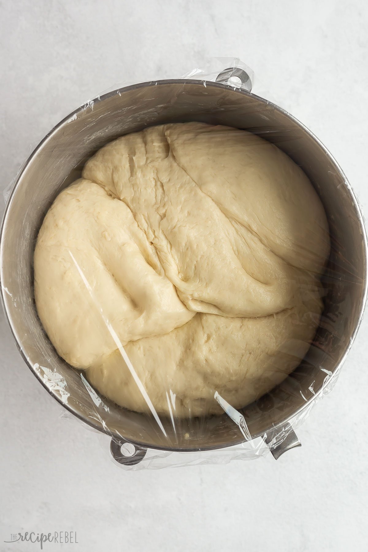 hamburger bun dough in steel mixing bowl with plastic wrap after rising