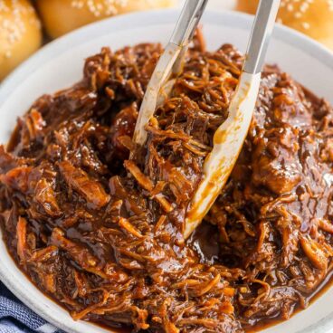 tongs picking up pulled pork from large bowl with buns in the background.