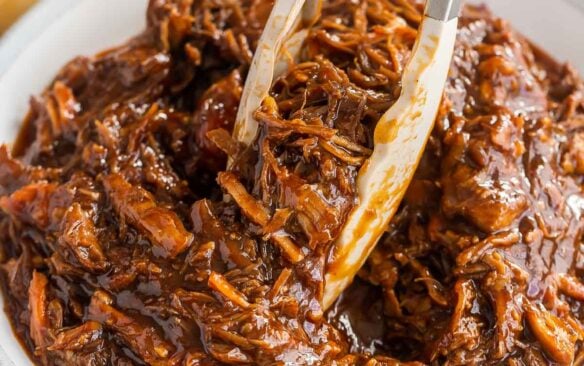 tongs picking up pulled pork from large bowl with buns in the background.