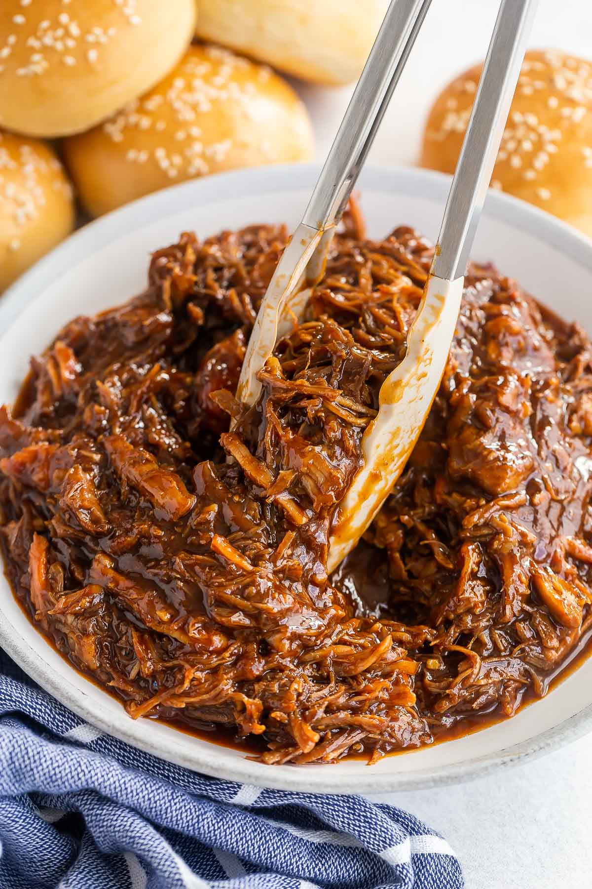 tongs picking up pulled pork from large bowl with buns in the background.