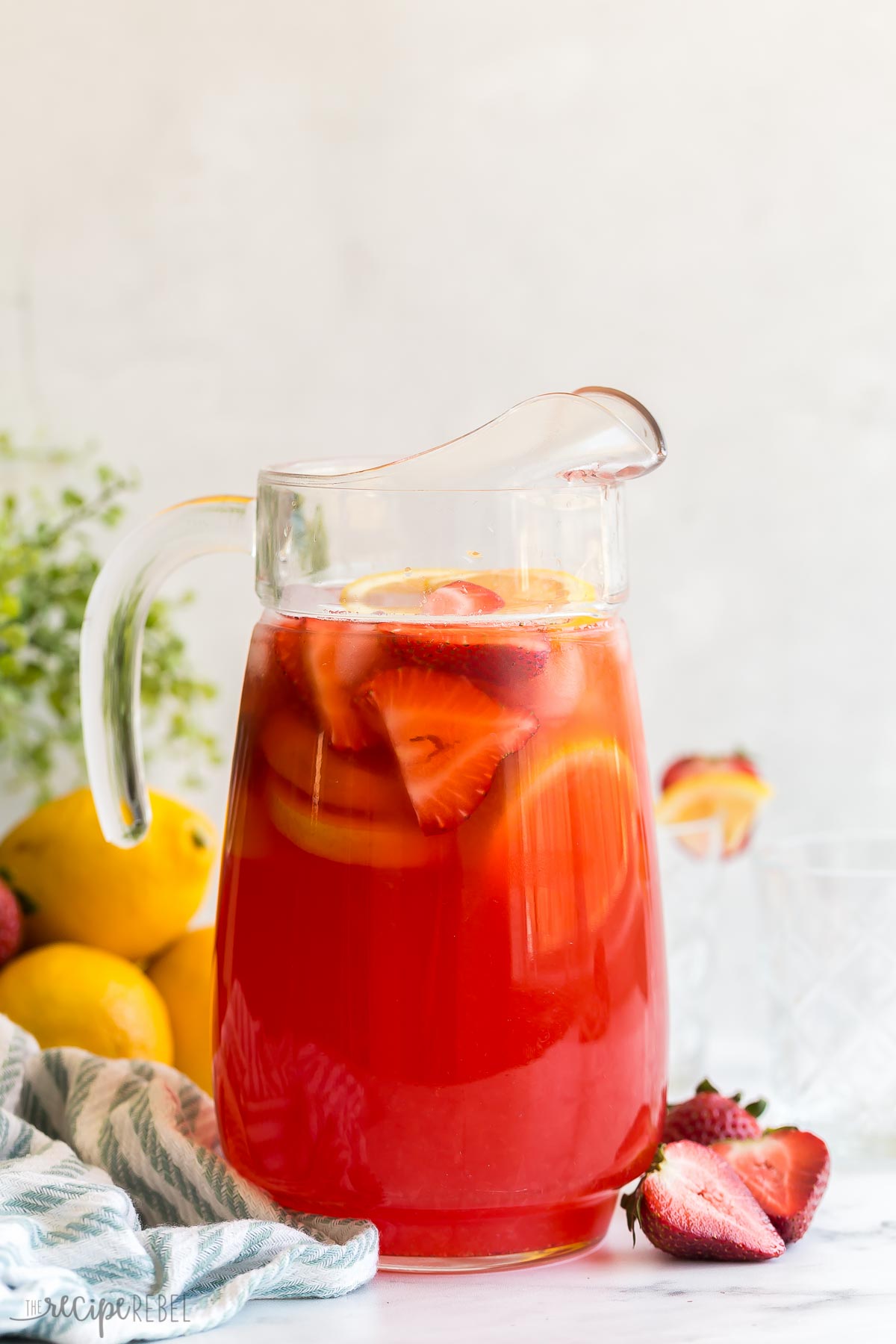 full glass pitcher of pink lemonade with lemons and strawberries.