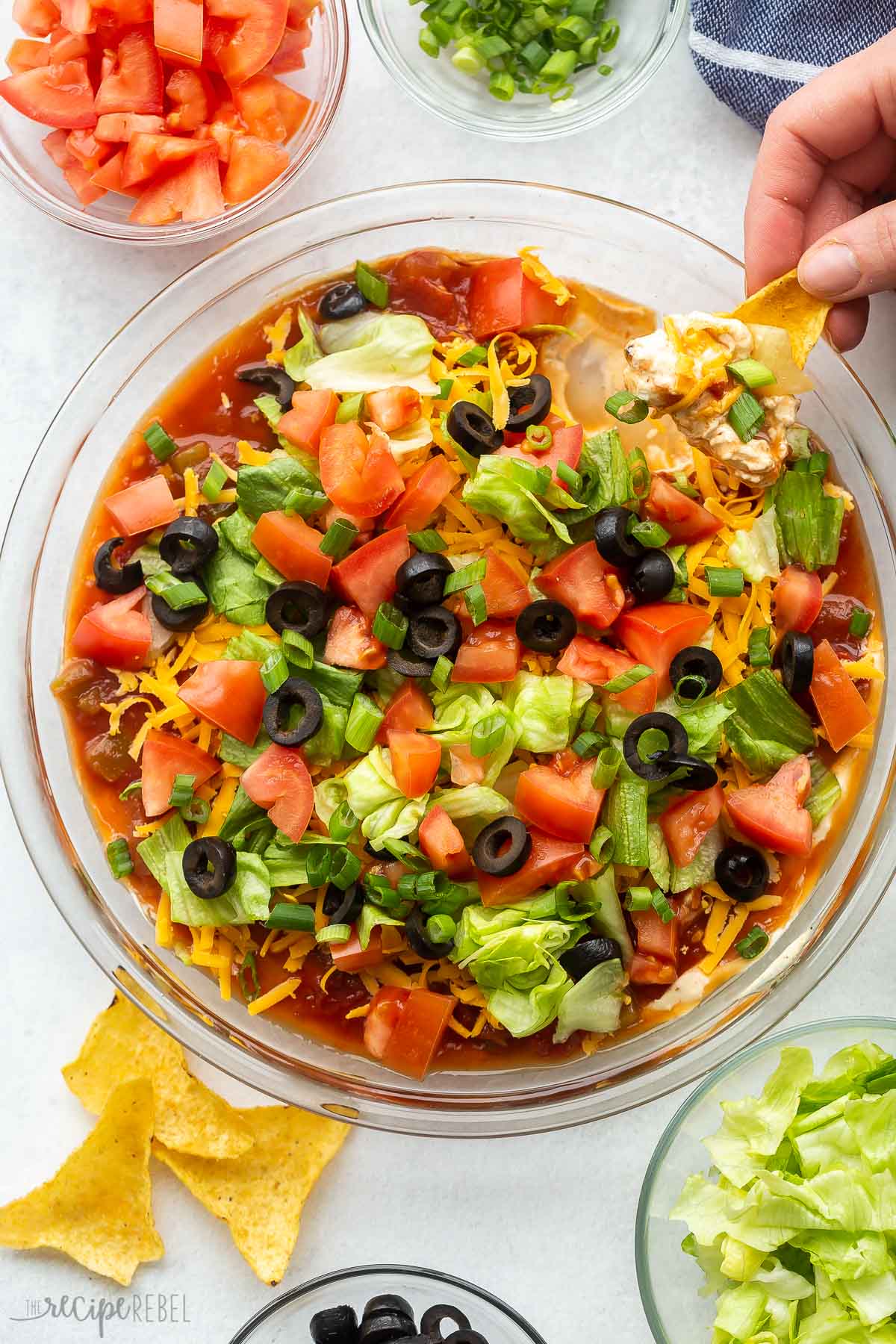 This Taco Dip is the perfect party appetizer. It's made up of a rich cream cheese base topped with the best taco fillings. Perfect served with tortilla chips! #tacos #dinner #recipe | appetizer recipe | appetizers | finger food | fun food | taco night | salsa | cream cheese | easy recipe overhead image of a hand scooping taco dip.