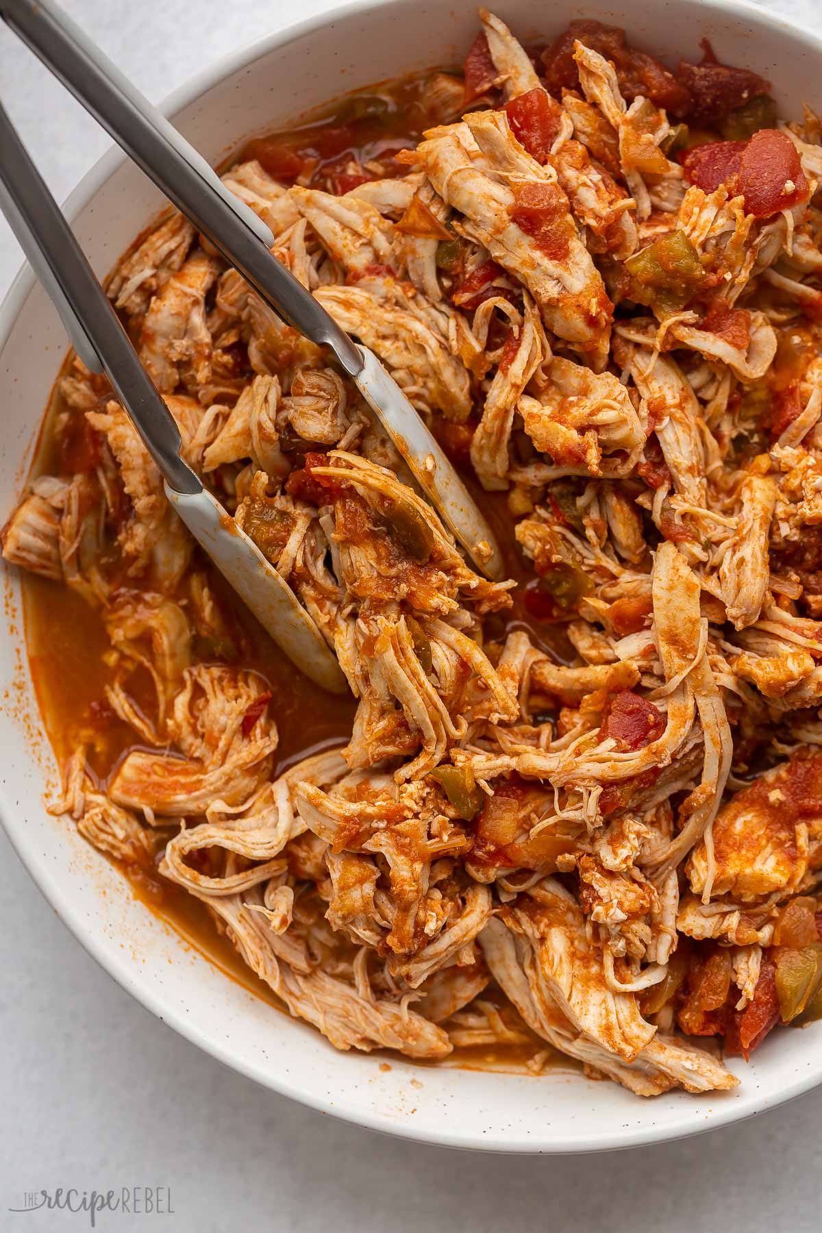 close up image of tongs grabbing instant pot salsa chicken from large bowl.