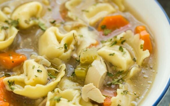 close up image of chicken tortellini soup in white bowl on grey background.