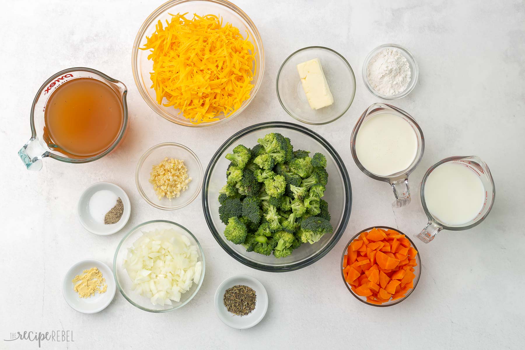 Top view of glass bowls of broccoli cheddar soup ingredients on grey background.