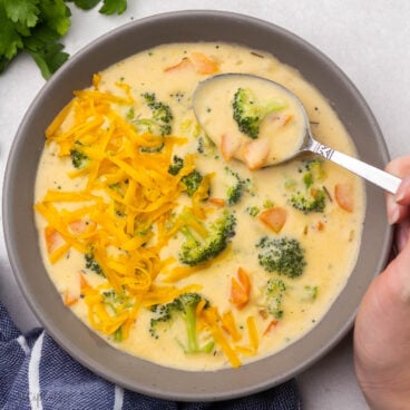 close up of a bowl of broccoli cheddar soup with a hand and spoon.