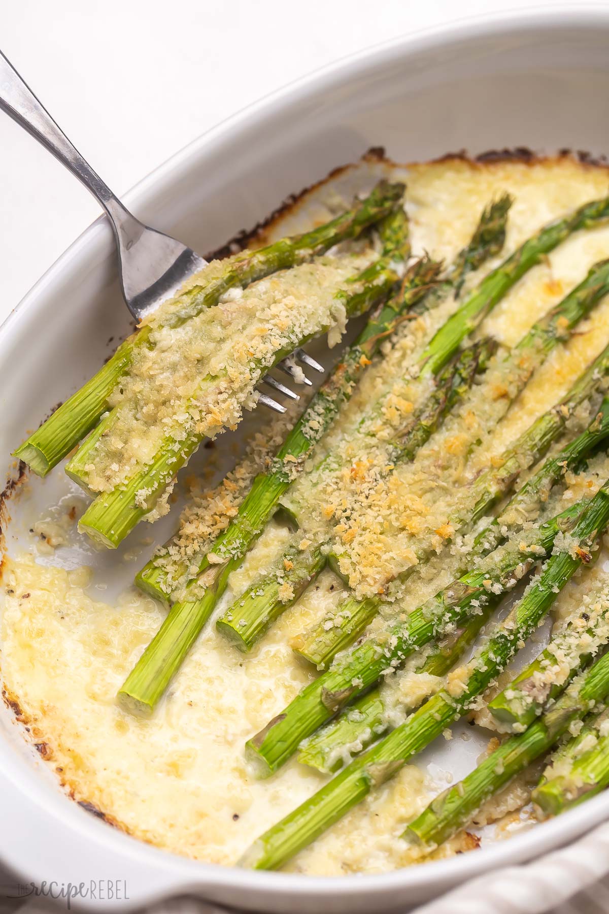 a fork scooping cheesy baked asparagus out of white baking dish.