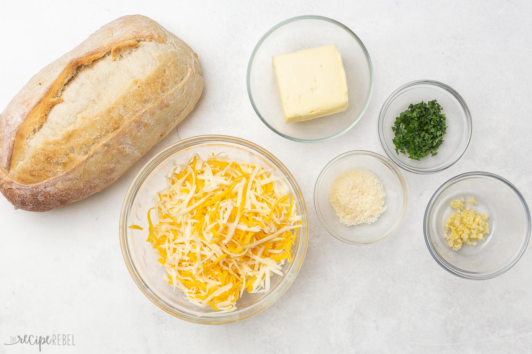 Top view of cheesy garlic bread ingredients in glass bowls on grey surface.