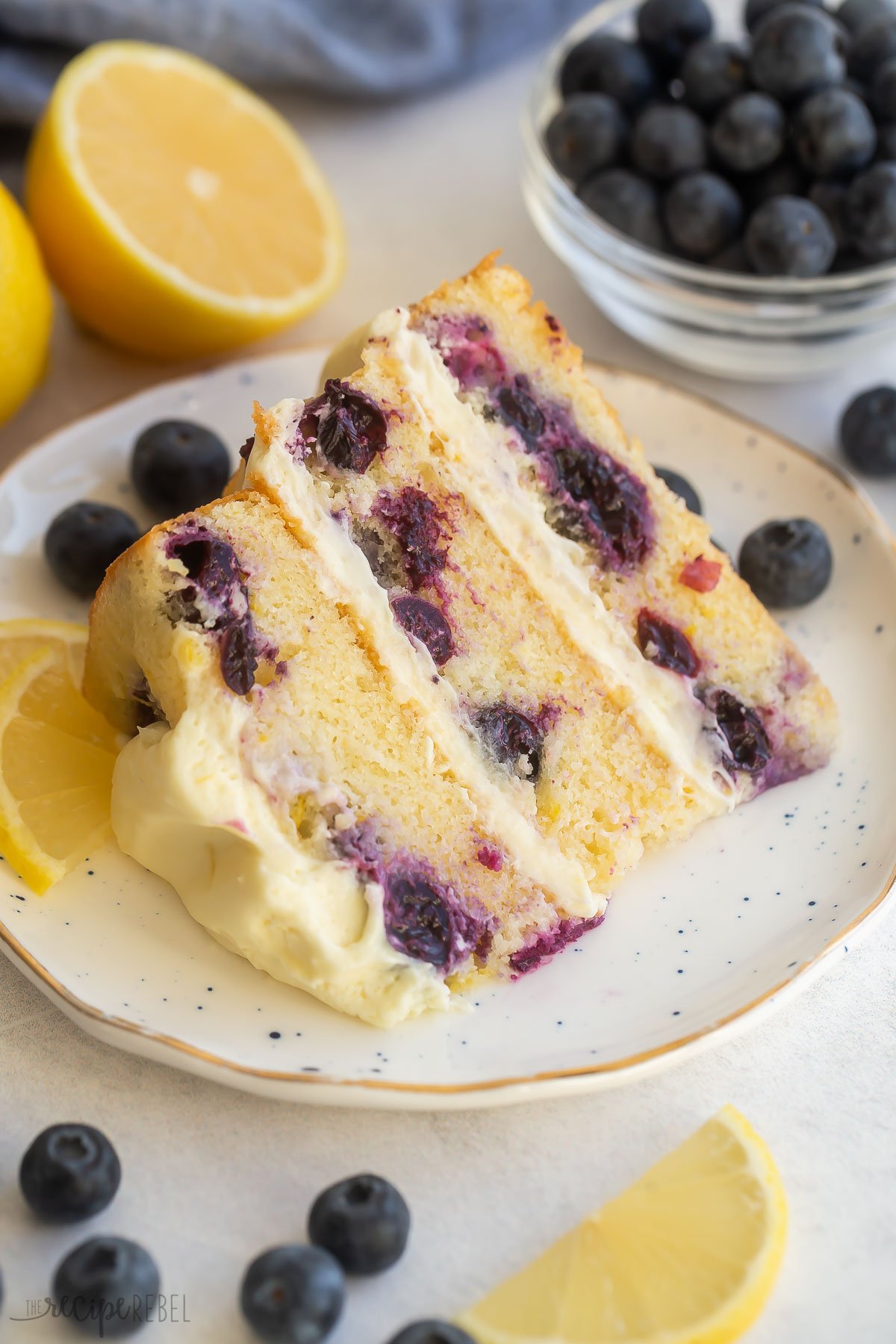 Close up of a slice of lemon blueberry cake on white plate with lemons and blueberries around it.