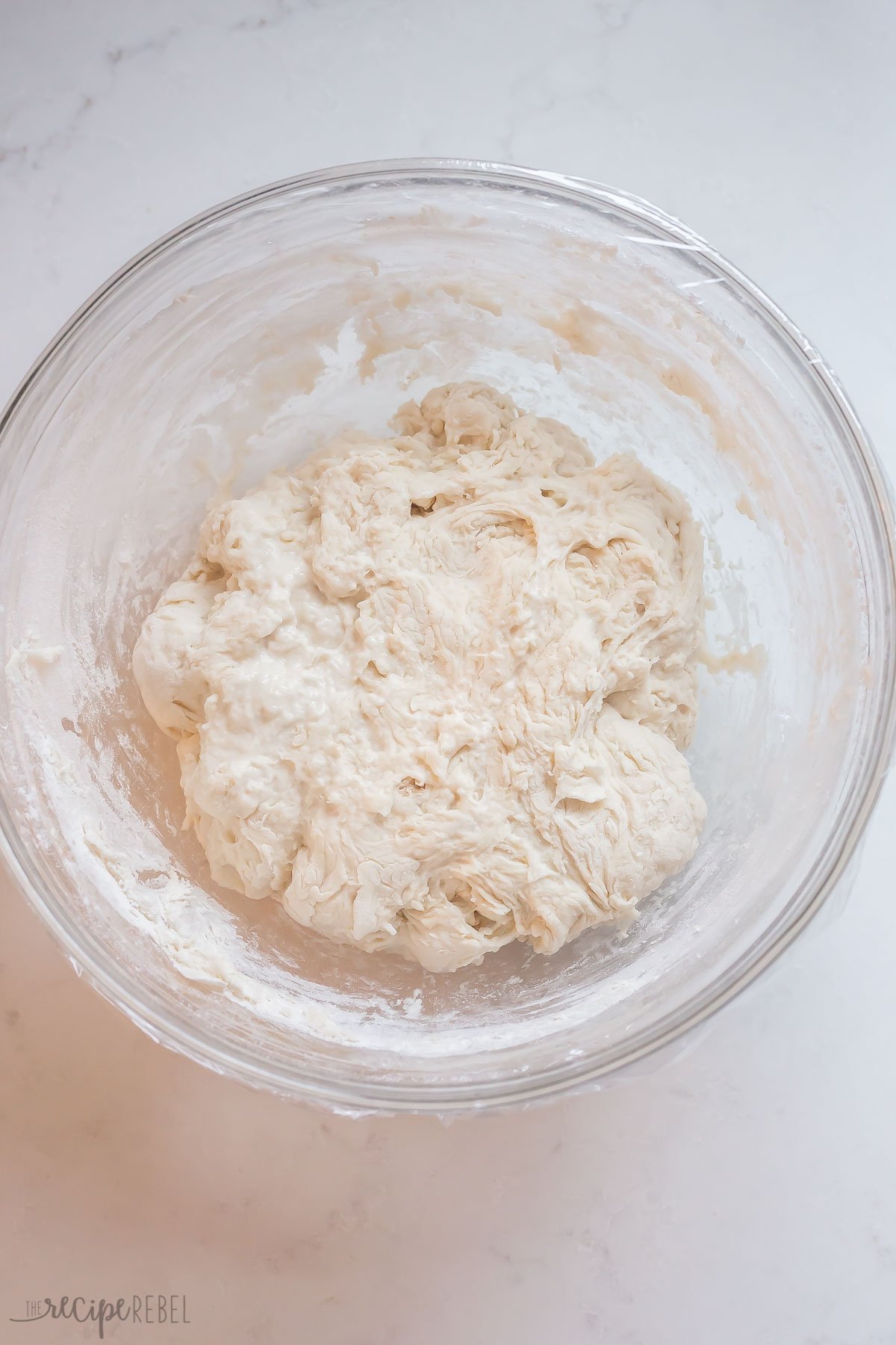 mixed no knead bread ingredients in glass bowl with plastic wrap on top.