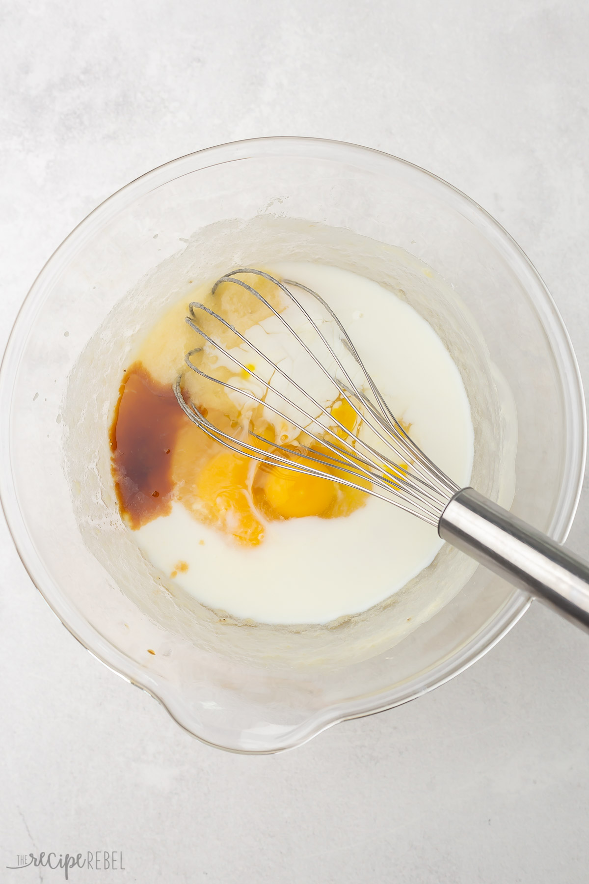 Top view of a glass mixing bowl with eggs, milk and vanilla extract on top of a banana mixture and a whick in it. 