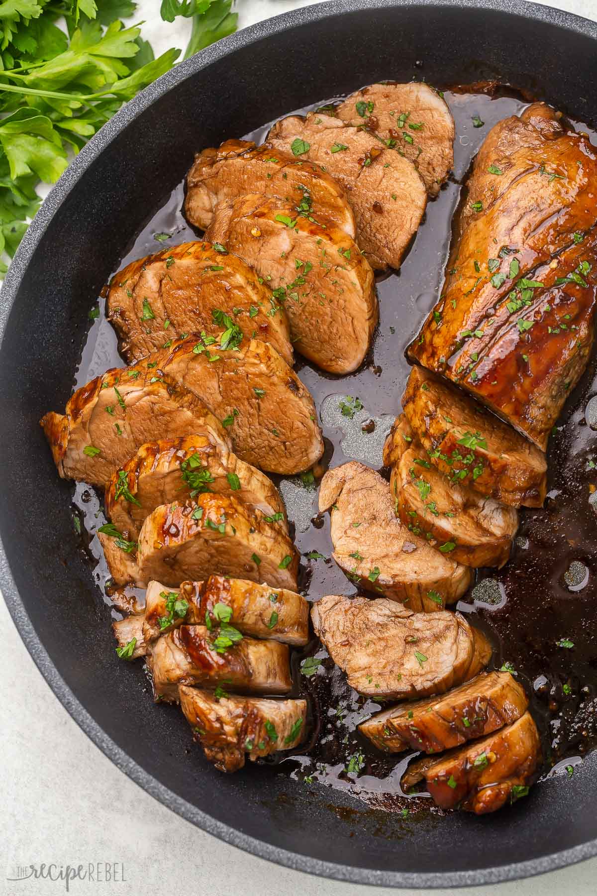 Top view of sliced pork tenderloin in black pan with parsley garnish.