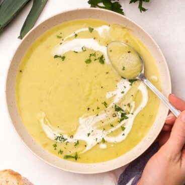 A spoon scooping potato leek soup out of a bowl with leeks and bread lying beside it.