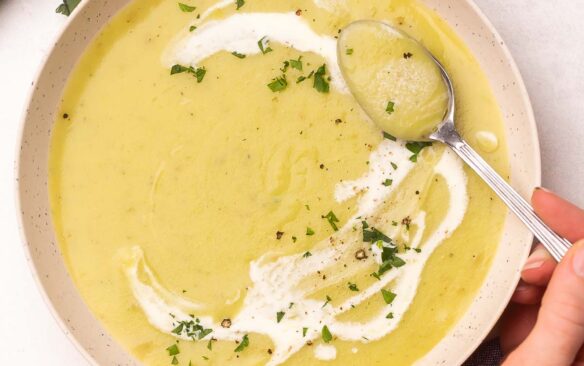 A spoon scooping potato leek soup out of a bowl with leeks and bread lying beside it.