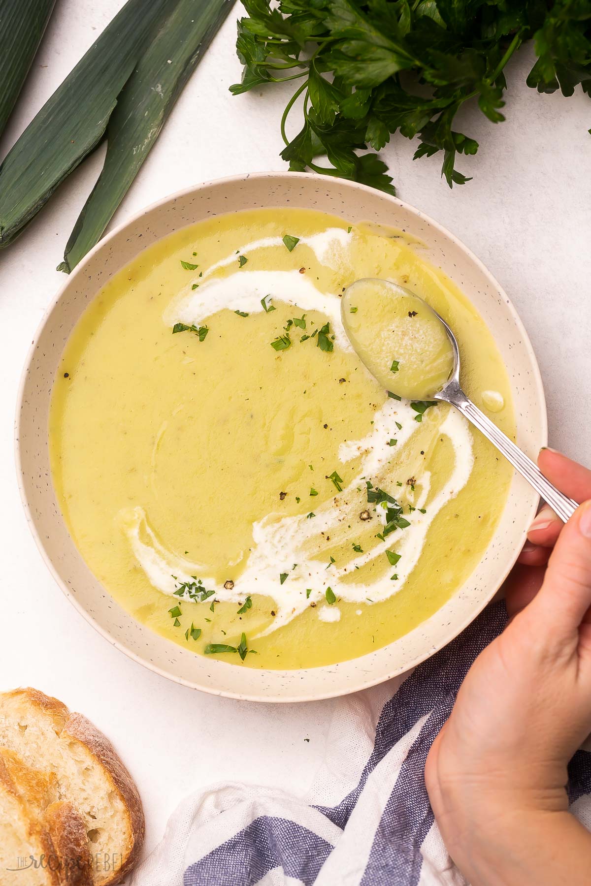 A spoon scooping potato leek soup out of a bowl with leeks and bread lying beside it.