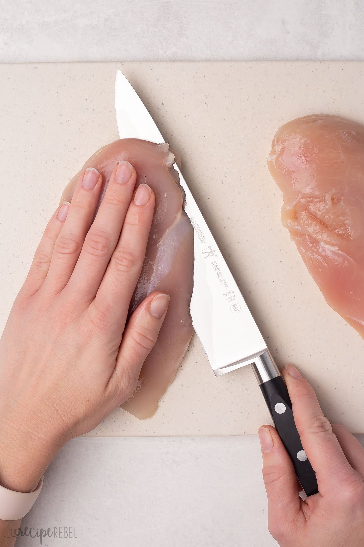 top view of a chicken breast being sliced in half on a white cutting board.